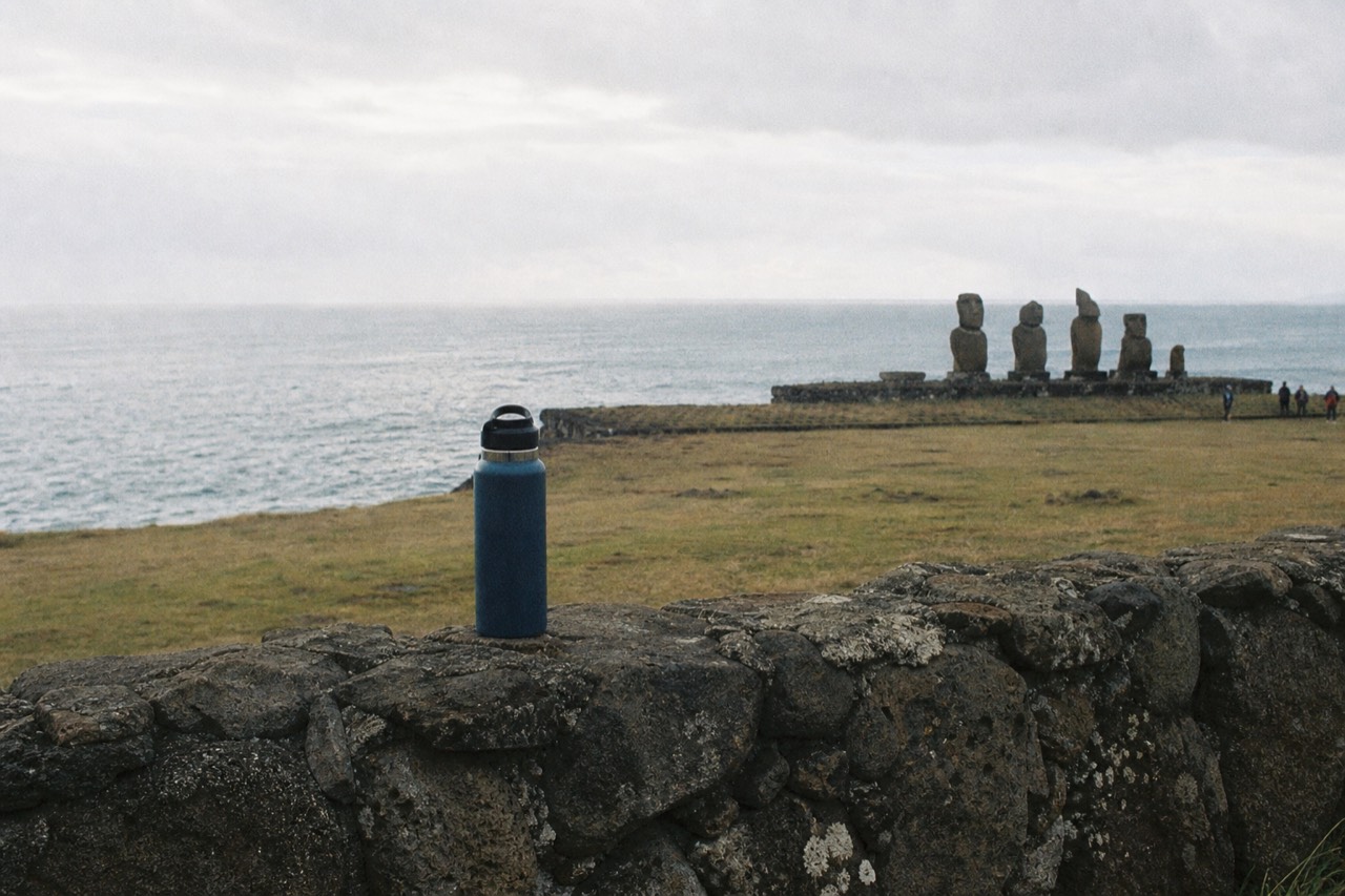 Reusable metal water bottle standing on a low stone wall above a bare grassy headland with the ocean stretching out behind it