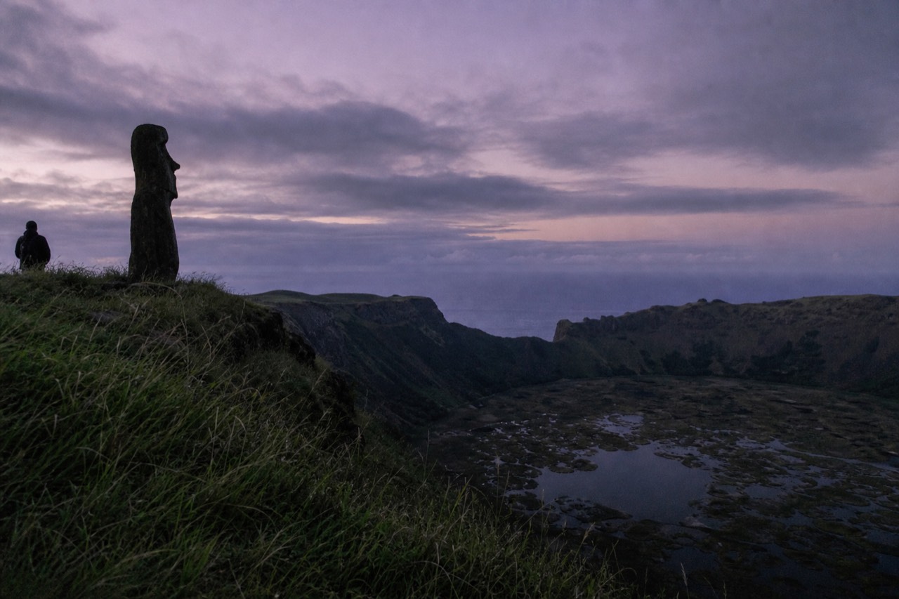 Steep grass-covered crater rim at Rano Kau dropping away to a reed-covered lake far below with the ocean beyond the far edge
