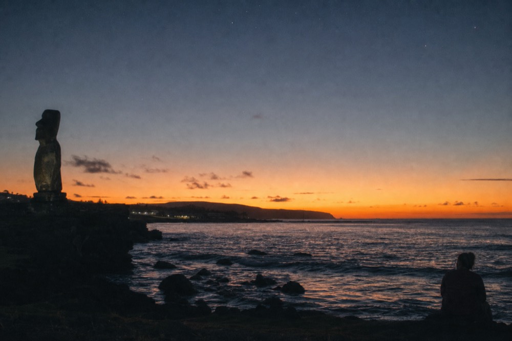 Stone ahu at Tahai silhouetted against an orange sunset with the Hanga Roa shoreline in the foreground