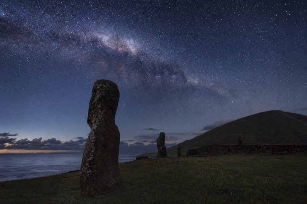 Night sky thick with stars and the Milky Way arching over a dark grassy headland near the coast