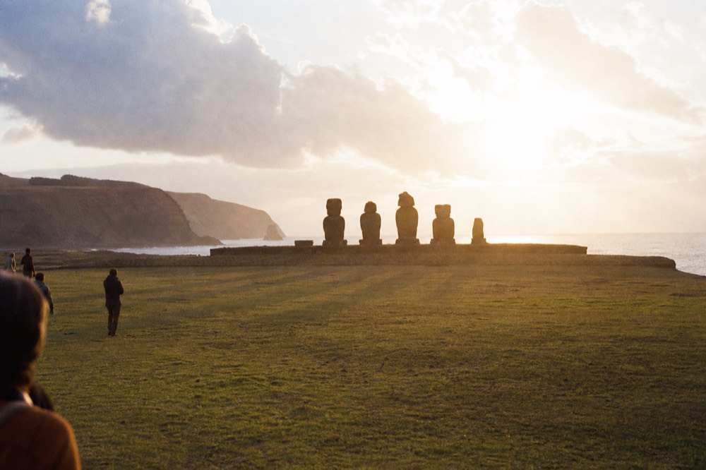 Seven stone figures on a low inland platform seen across open grassland with distant hills behind