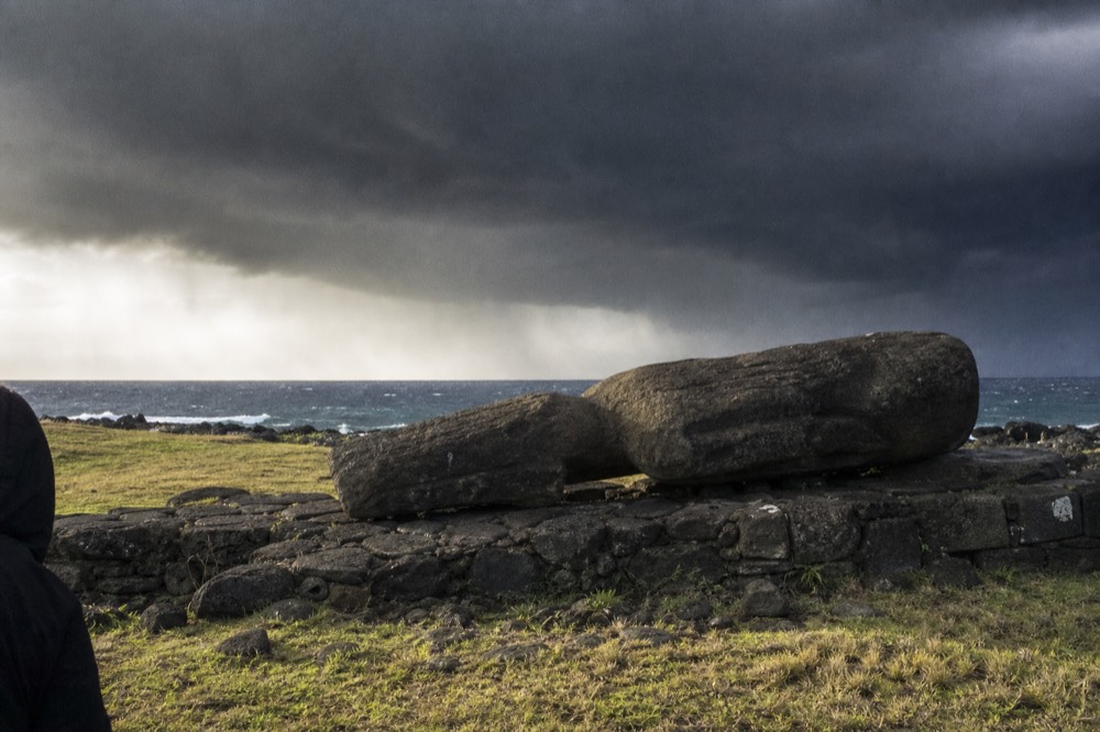 Fallen moai lying face down on broken platform stones with the ocean beyond a low rocky shoreline